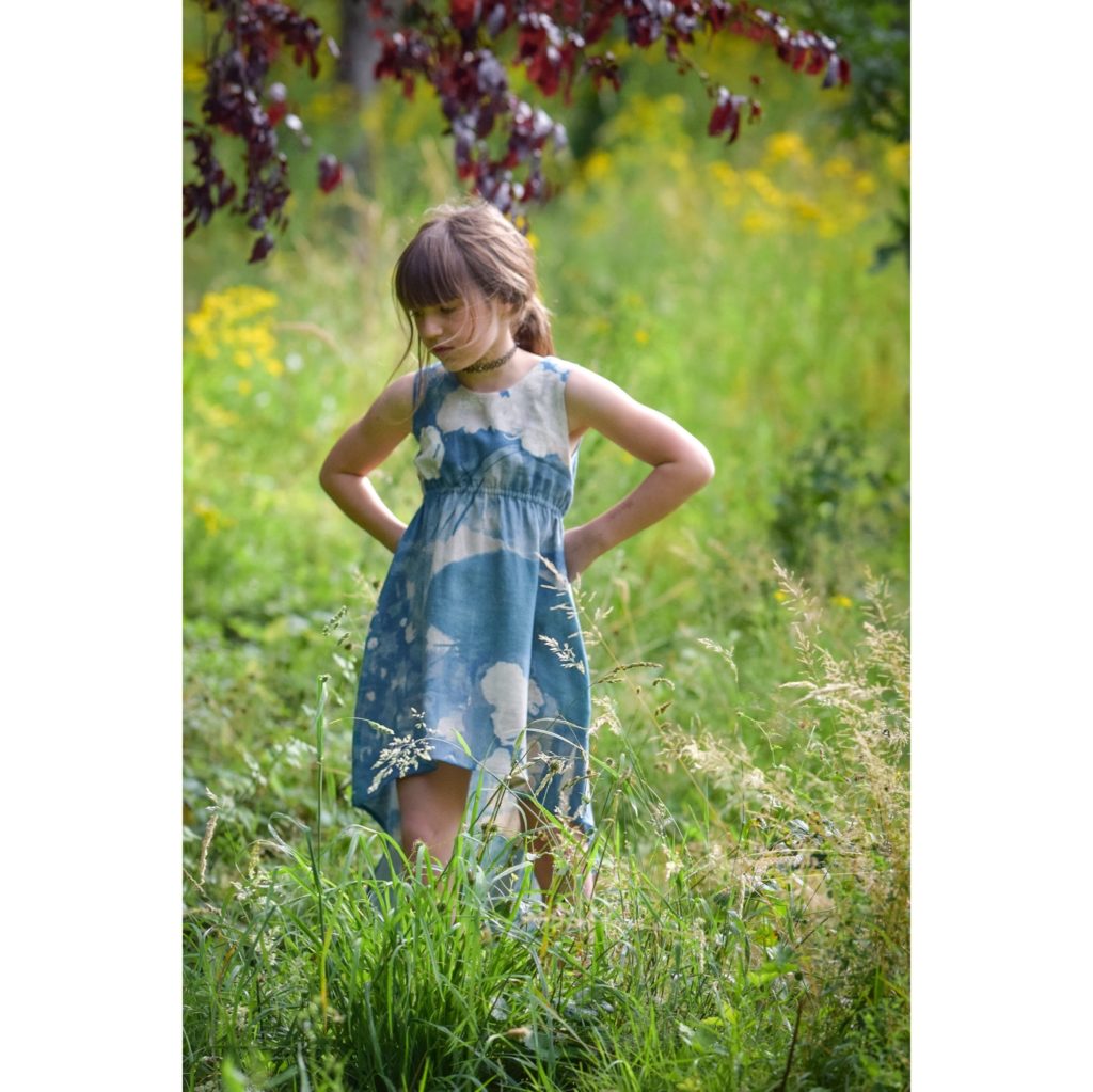 Robe asymétrique cintrée bleue, à motif partiellement floral, sans manche, portée par une petite fille se tenant debout dans l'herbe.